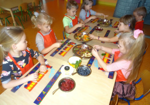 Grupa dzieci siedzi przy stole w ręku trzymają listki mięty, które wrzucają do misek wypełnionych owocami.